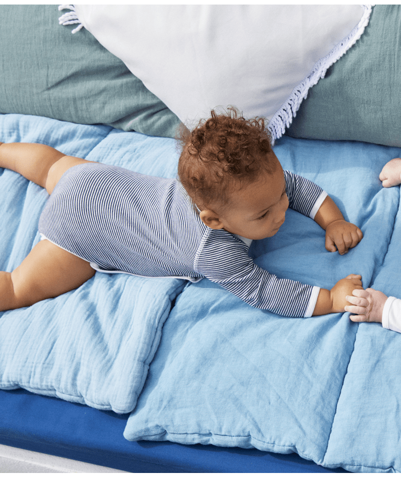 Bébé allongé sur un lit bleu, portant un body rayé, tendant la main dans une scène douce et naturelle du quotidien.
