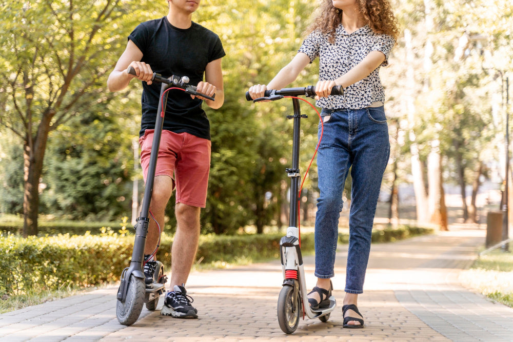 Deux adultes côte à côte sur des trottinettes dans un parc, illustrant l'utilisation de trottinettes en milieu urbain.