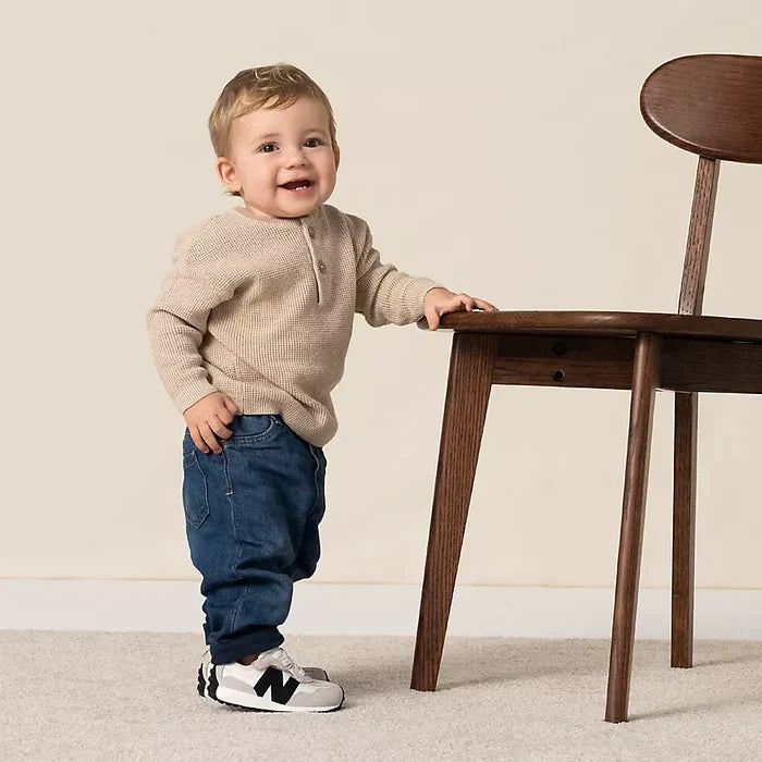  Bébé garçon debout portant des baskets New Balance blanches et noires, un pull beige en maille et un jean bleu