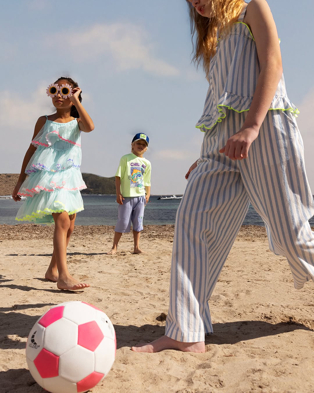 Trois enfants jouant au football sur la plage en tenues d'été colorées.