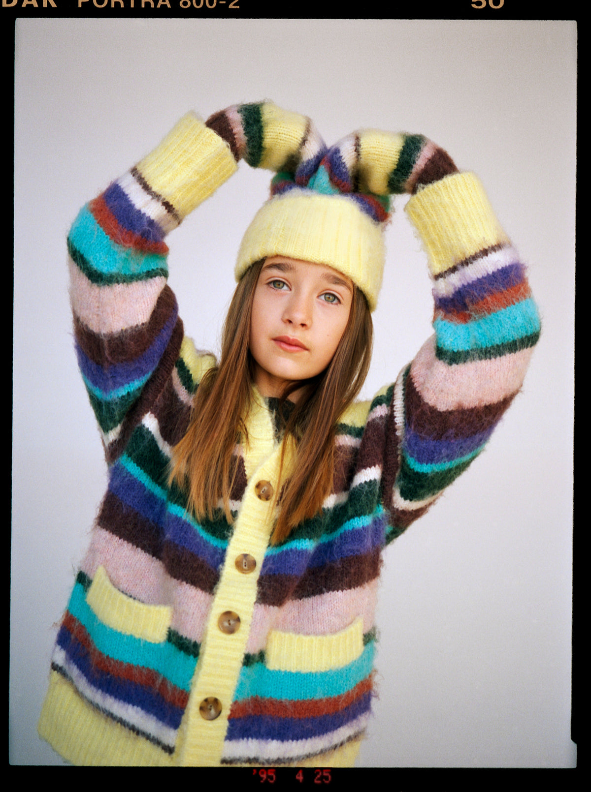 Enfant portant un cardigan Molo rayé multicolore et une tuque jaune, les bras levés au-dessus de la tête
