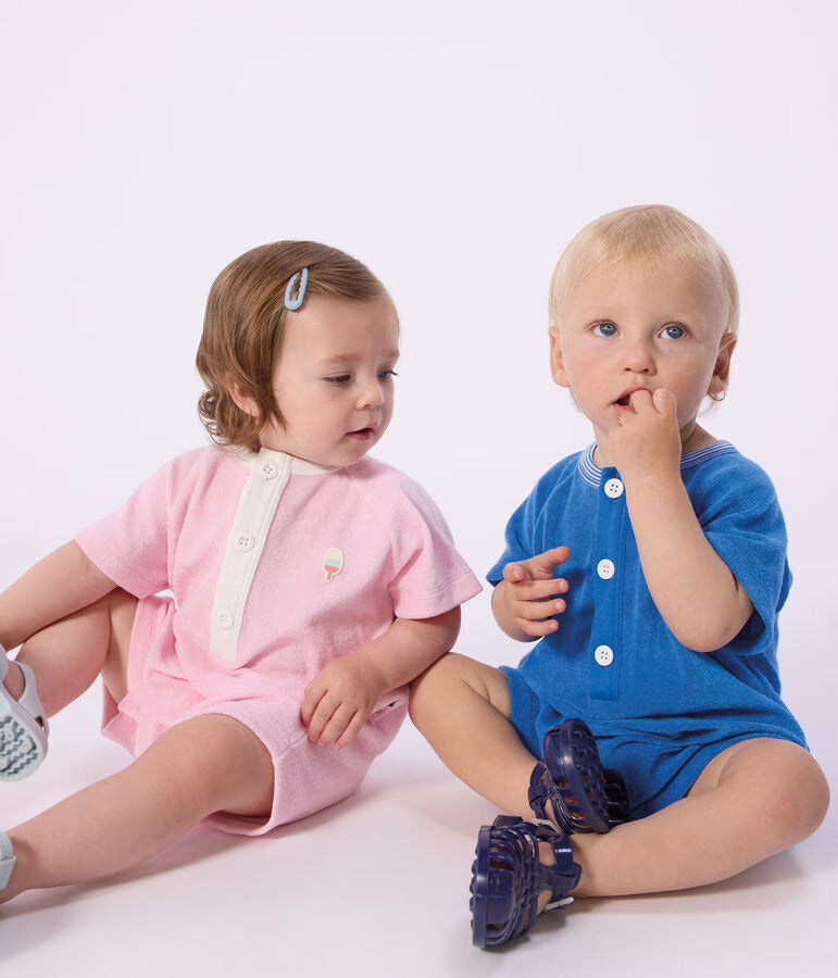 Deux bébés habillés en vêtements Petit Bateau enfants : barboteuse rose à broderie glace et barboteuse bleue à boutons blancs, assis côte à côte