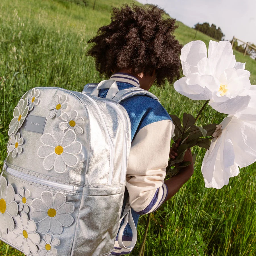 Garçon dans un champs, portant un sac à dos argenté avec des marguerites en 3D sur l'ensemble.